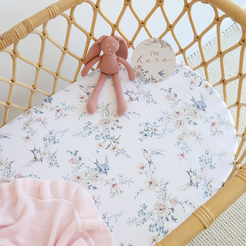 Floral crib sheet with a pink plush toy and 'I'm here' sign in a wicker crib.