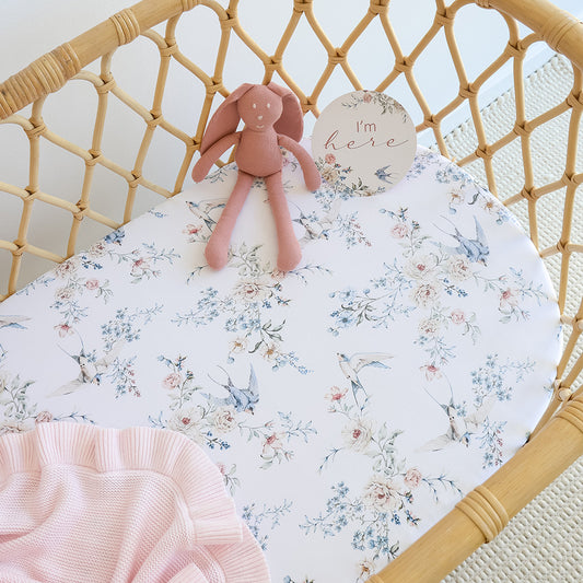 Floral crib sheet with a pink plush toy and 'I'm here' sign in a wicker crib.