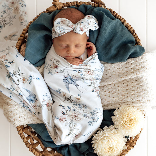 Newborn baby swaddled in a floral blanket with a bow headband, lying in a woven basket.